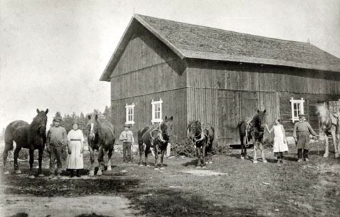 Människor och hästar som står framfr en ladugårdsbyggnad.