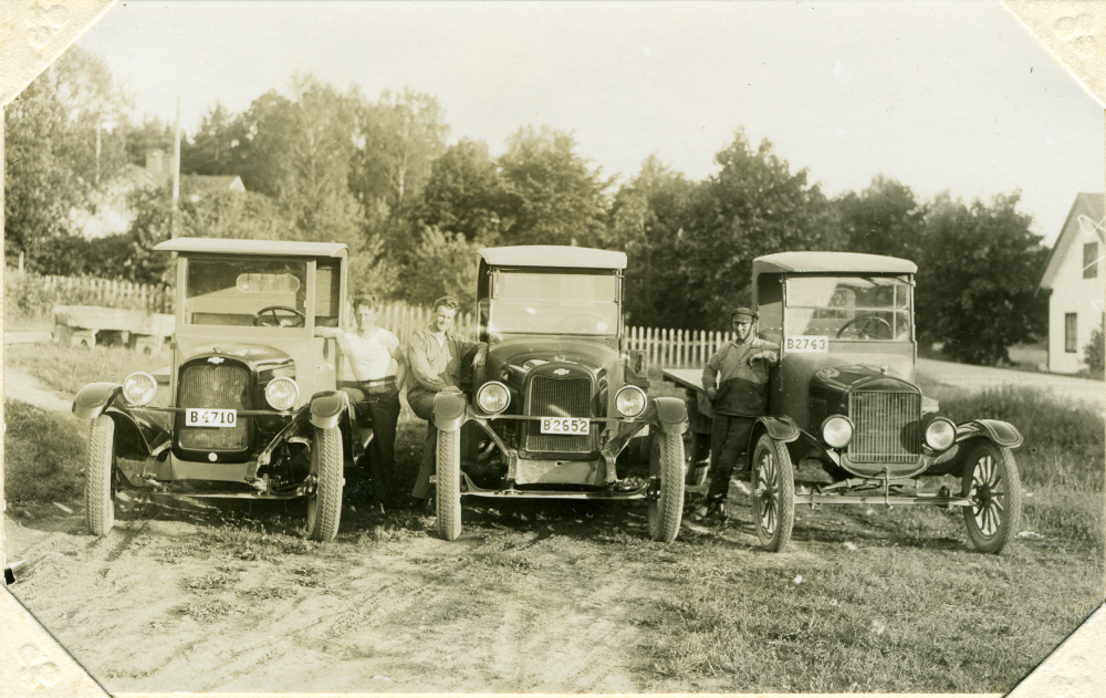 De tre bilarna, de två till vänster av märket Chevrolet, ägdes av bröderna Ingvald och Verner Gustafsson som hade ett åkeri. Ingvald står till vänster, Verner i mitten och till höger står brodern Ruben som var anställd hos dem. 
Bilden är tagen på andra sidan vägen räknat från Åbyholms gård, där bröderna bodde med sin mamma och flera syskon. På bilden syns gaveln till en av flyglarna. 
Runt 1933 flyttade Verner och Ingvald till Täby, nuvarande kyrkbyn, där de hade taxi- samt åkerirörelse. Ingvald hade hand om lastbilarna och körde mycket tegel från Vallentuna tegelbruk. Verner stod för taxirörelsen.
Original och uppgifter: Birgit Franzén