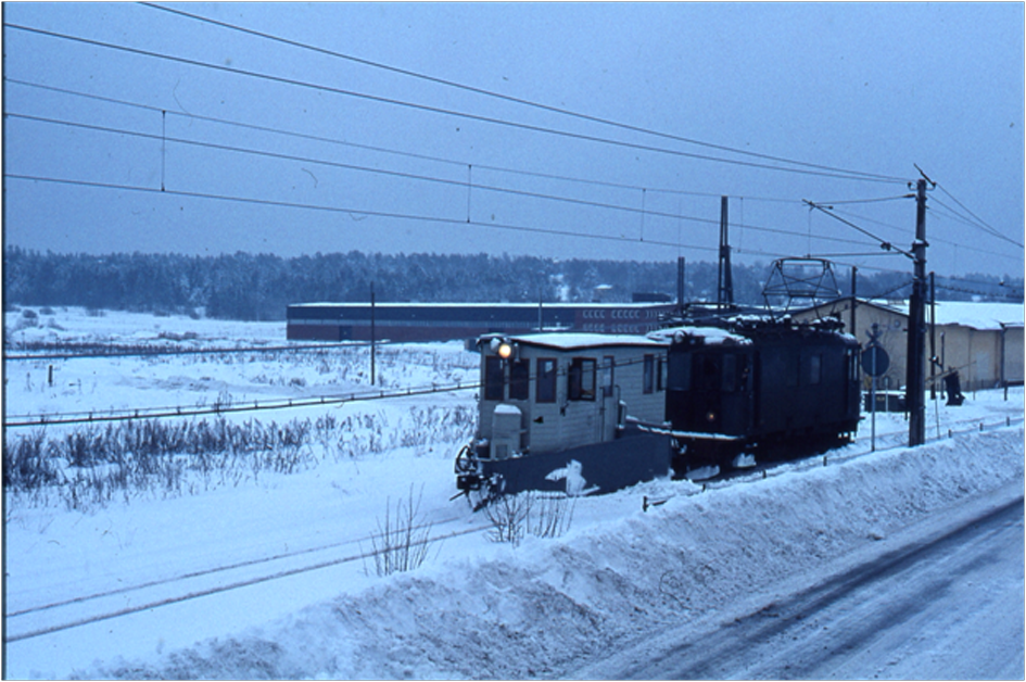 Snöplogen, påskjuten av lok 3255, på väg norrut från Vallentuna.  Tåget har just passerat Åby gata.  I bakgrunden ser man  motorvagnsstallet och t v  en ny utbyggnad vid Tellus (nu Vallentuna sporthall). Banvägen t.h.