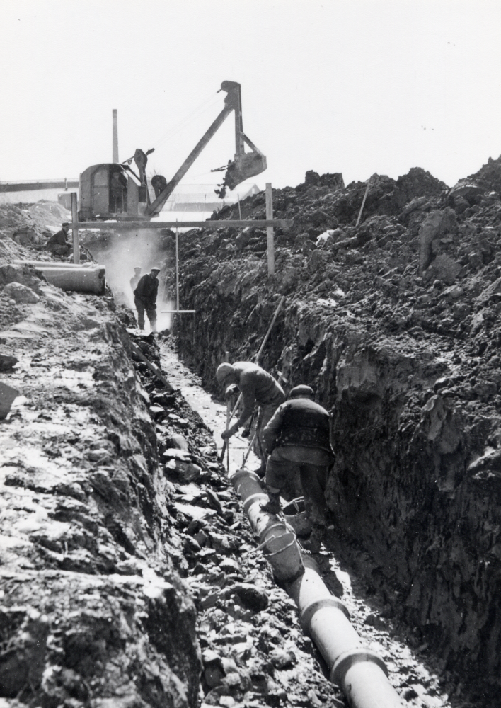 Arbeten med nya ledningar för VVS i Ormsta. En grävskopa har grävt djupa schakt, där män lägger ner avloppsrör.  I bakgrunden syns Åby tegelbruk