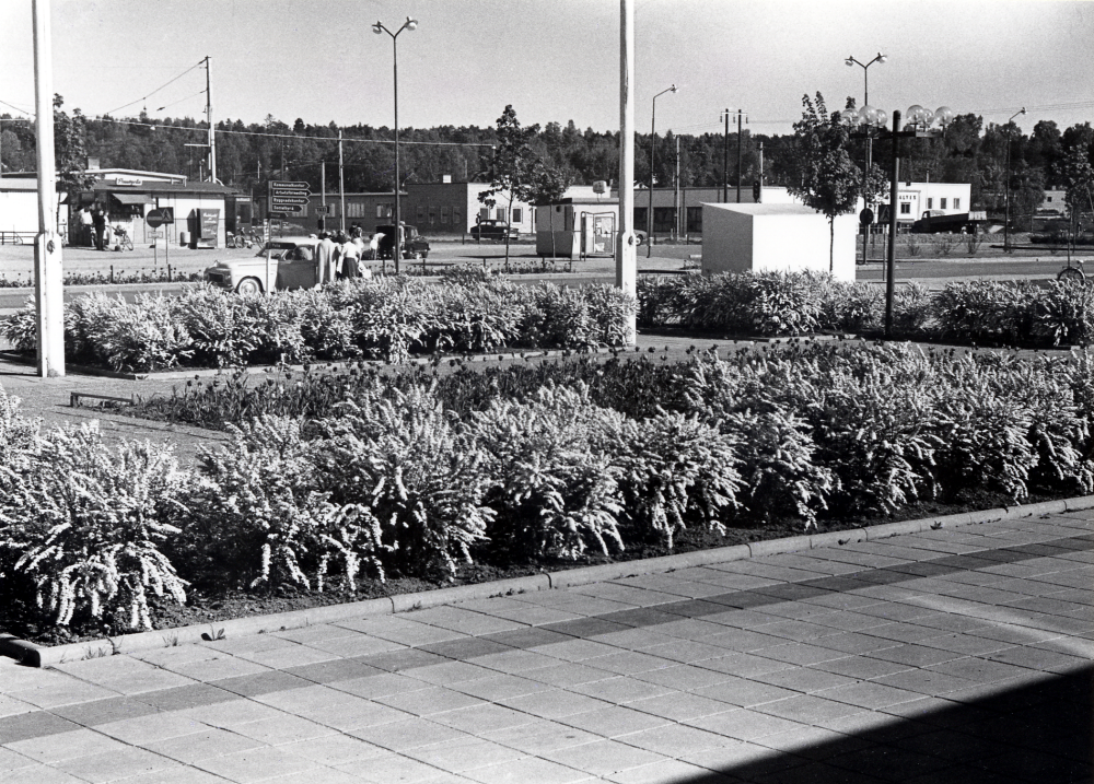 Planen framför Vallentuna kommunalhus. I förgrunden planteringar, Banvägen och planen utanför Vallentuna järnvägsstation där man fr v ser Pressbyråns kiosk och en korvkiosk. På andra sidan järnvägen syns taket på K S Karlssons smedja, Elverket och Vallentuna motor, alla med adress Industrivägen