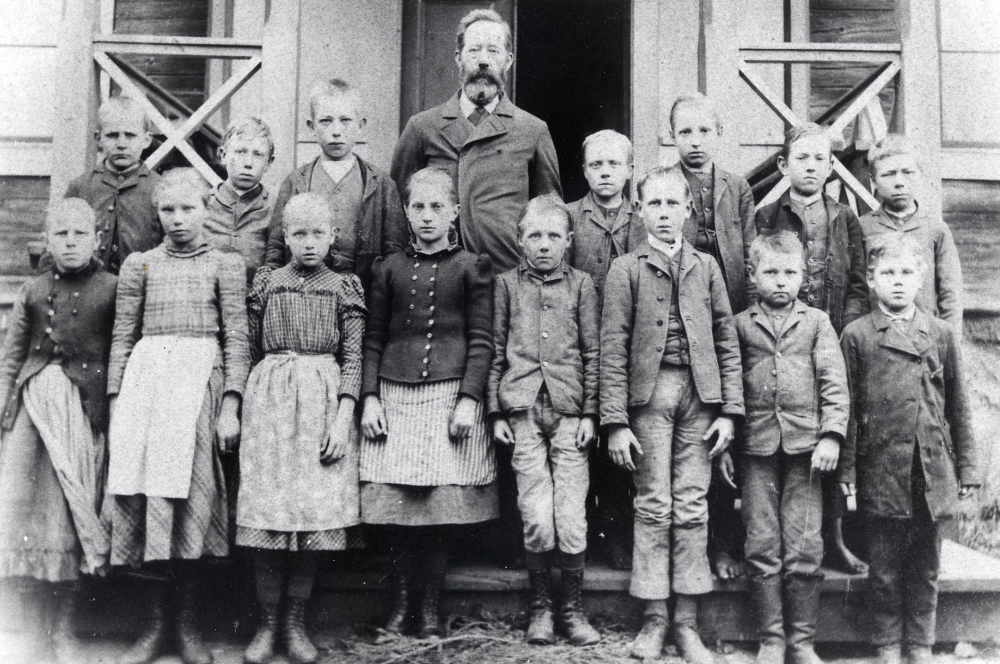 Förmodligen Angarns skola. Lärare förmodligen C Fromm, far till läraren i Vada Brynolf Fromm. Framför läraren står Nanny Skoglund född 1883, som sedan blev lärarinna i Kyrkskolan i Össeby-Garn.
Uppgift från Riksarkivet: Nanny Vilhelmina Skoglund föddes 1883 i Lilla Garn, Össeby Garn.