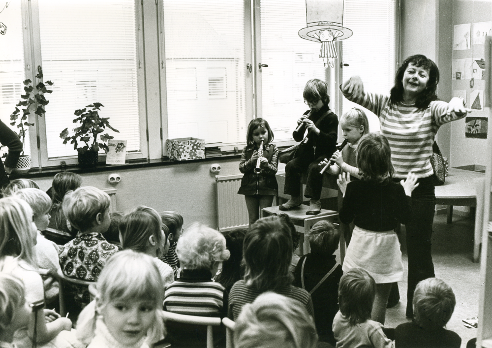 Skådespelaren Elisaveta leker med en grupp barn på Vallentuna bibliotek, tidigt 1970-tal. Tre barn spelar flöjt / blåsinstrument.
•Fotografiet ingår i bildserien ”Vardagsliv i Vallentuna kommun” av Rachael Gough-Azmier & Gunilla Blomé, Haga Studios HB.
