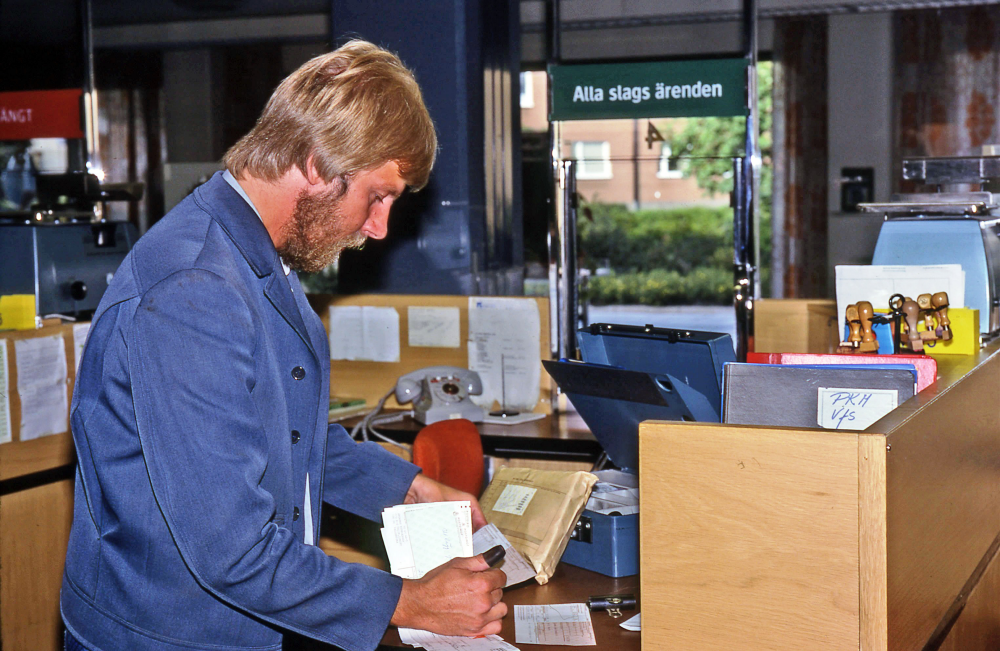 En arbetsdag med lantbrevbärare Mikael Mattsson. Postkontoret på Tuna torg; Lantbrevbärare Mikael Mattsson hämtar pengar, paket och rekommenderade brev på postkontoret på Tuna torg. Här går han igenom avier för att se hur mycket pengar som behövs under turen. Vissa turer kunde brevbärarna ha upp emot 100.000 kr med sig. Ett sedelkuvert står lutat mot kassaskrinet. Genom fönstret i bakgrunden syns Gärdesvägen.
