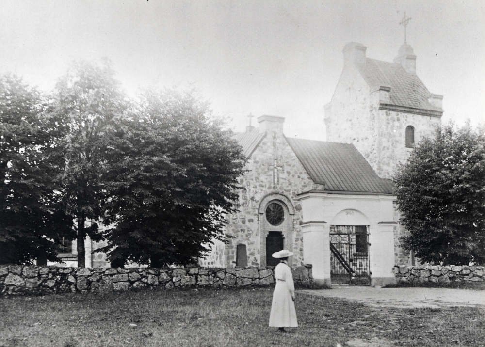Vallentuna kyrka. En kvinna i vit hatt står på planen utanför kyrkgrindarna. Förmodat motivdatum ca 1900-1910