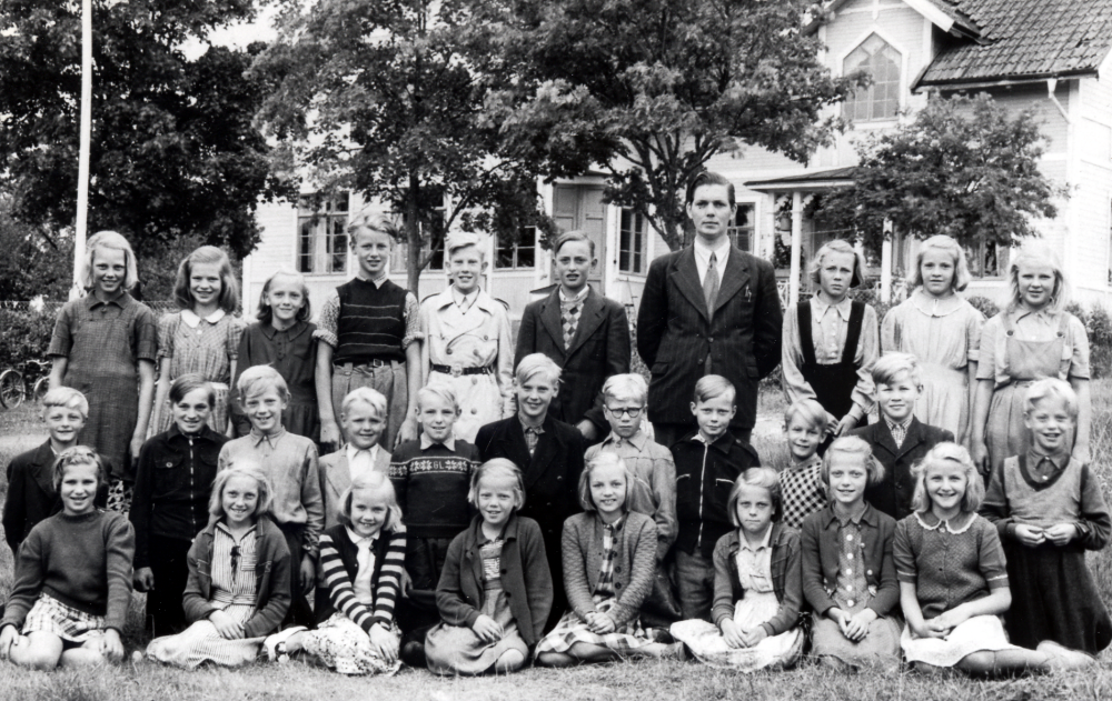 Läraren Gösta Abrahamsson med elever utanför Vada skola.  I övre raden står fr v Svea Jansson, Gun Jakobsson, Ulla Jonsson, Rune Alstermark, Klas Bergman, Ingvar Flodin, Kerstin Gustafsson, Britt Karlsson och Greta Mokvist.  Andra raden fr v Sven-Olov Viman?, Olle Andersson, Sten Sjögren, Lasse Mokvist, Gunnar Söderström, Kalle Mokvist, Lennart Gustavsson, Ingvar Gustavsson, Karl-Gustav Adegren och Göte Sjögren (tvilling med Sonja).  Sittande fr v Kerstin Wallerts?, Edit Jansson, Kerstin Alstermark, Sonja Sjögren (tvilling med Göte), Gunvor Eriksson, Barbro Pettersson, Ingegärd Sjögren och Kerstin Törnblom