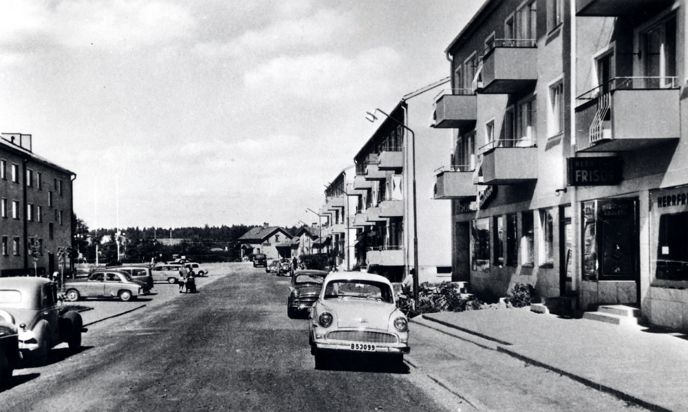 Centralvägen, Vallentunas första affärsgata, med flerfamiljshus och affärslokaler som byggdes i början av 1950-talet. Till höger finns en frisersalong, skylten visar att det är både herr- och damfrisör. Bredvid ligger Konsum snabbköp som senare blev lokal för Vallentuna varuhus. I bakgrunden mellan husen skymtar Vallentuna järnvägsstation med stationshuset till höger och till vänster järnvägsrestaurangen Flugan. En Opel Rekord står parkerad på Centralvägens högra sida (enligt uppgiftslämnaren kom bilmodellen ut 1956 i denna version). På andra sidan går Elsie Westlund med barnen Lars och Tommy och Kjell i barnvagnen.
(En bättre fotokopia finns, se löpnr 8)