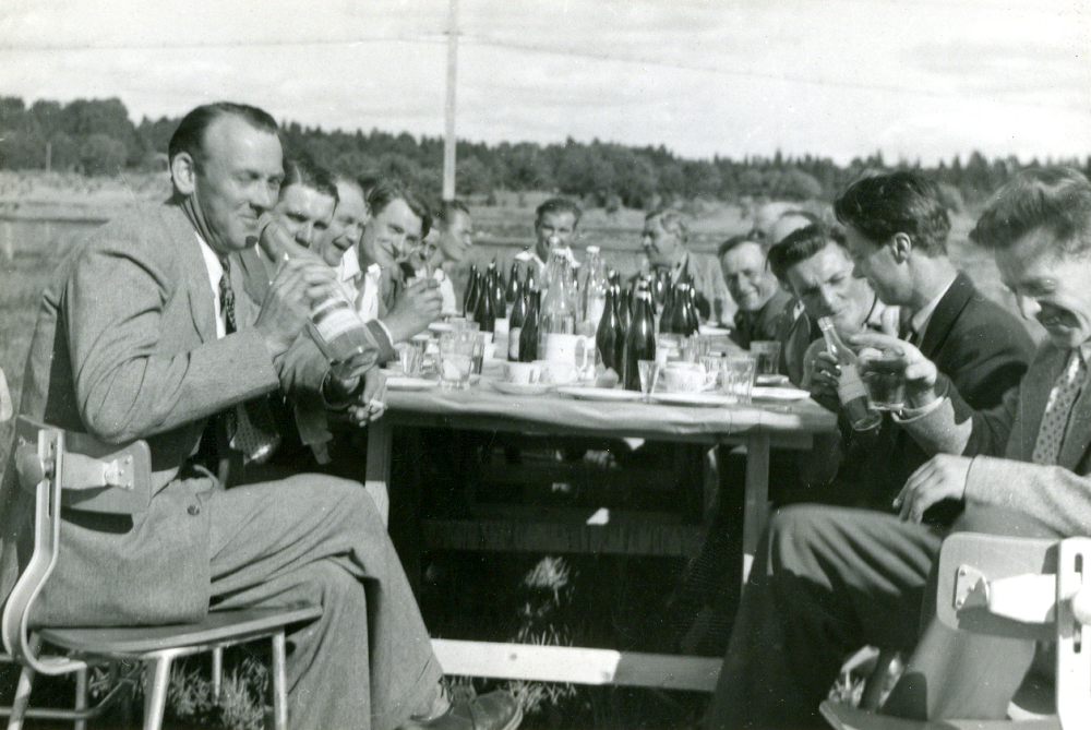 Utanför Vallentuna möbelindustri vid Banvägen har personalen taklagsfest . Man sitter utomhus utanför fabriken med Roslagsbanans spår i bakgrunden. Rya Idermark och ägde fabriken tillsammans med Ingvar Johansson.  Runt bordet sitter på vänster sida Kalle Jonsson "Klot-Johan", Bertil Tillborg, resten okända, längst ner Rya Idermark.  Vid nederkanten t h Mörk.  På andra siden bordet närmast kameran sitter Bengt Edblom, ev Klintberg, Henry Bergqvist, resten sudda och okända.  Vallentuna möbelindustri tillverkade trähöljen till Luxor TV-apparater och och träfodral till radioapparater.