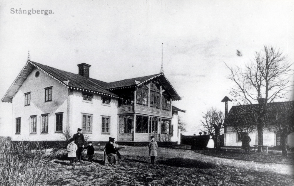 Stångberga gård. På flygelbyggnaden finns en vällingklocka. På planen t v om mangårdsbyggnaden står en man med två små barn, en kvinna sitter på stol med ett litet barn i knäet och en pojke står vid sidan om henne. Vid flygelbyggnaden står 3 svartklädda personer, en med vitt förkläde, förmodligen tjänstefolk, samt en man under det stora trädet