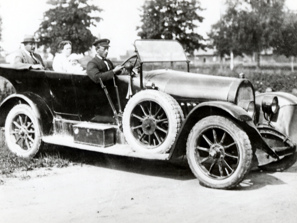 Personbil av 1920-års modell. Vid ratten sitter en uniforsmsklädd chaufför. I baksätet sitter en man och kvinna med ett litet barn.