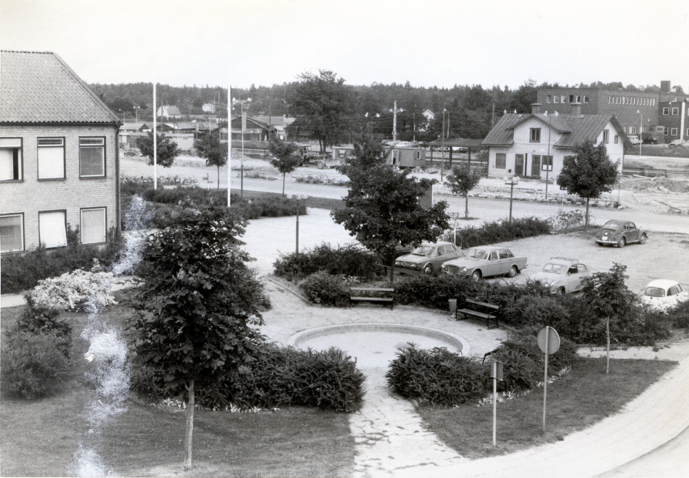 Vallentuna centrum tidigt 1970-tal, före augusti 1972. Parken vid gamla kommunalhuset med en liten damm, planteringar och parkbänkar i förgrunden, samt en bilparkering vid Skördevägen. Södra flygeln på gamla kommunalhuset syns till vänster. Vallentuna station och stationshuset syns till höger, på andra sidan Banvägen. I bakgrunden skymtar fabriken Tellus Maskin AB.
