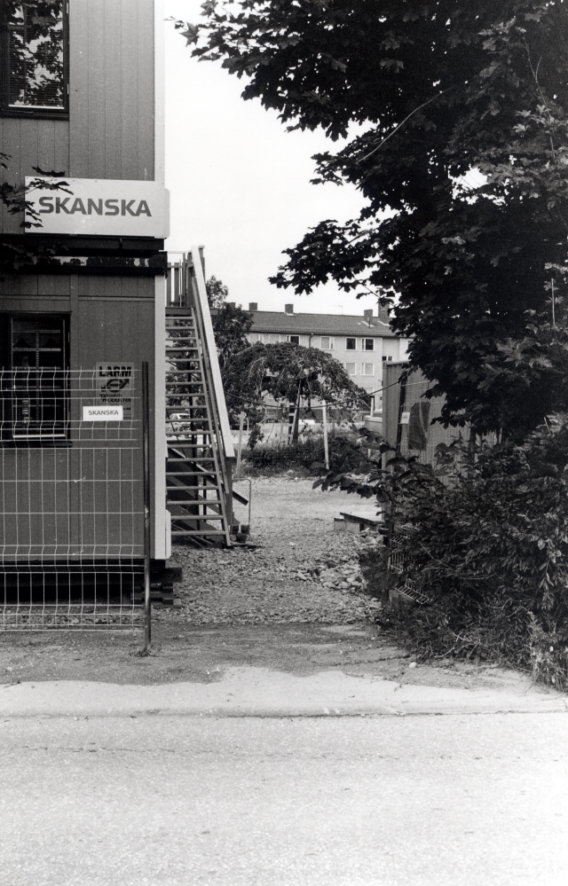 Byggarbetsplatsen i de s k Trädgårdskvarteren vid Mörbyvägen.  Byggherre är Skanska.  Områdets 54 bostadsrätter kallas Runhällen.  Här skymtar de äldre bostadshusen vid Sommarvägen mellan byggbarackerna sett från Mörbyvägen.  I bildens mitt syns den hängalm som tidigare växte i K E Karlssons trädgård, flyttat längre västerut i samband med byggnation av området