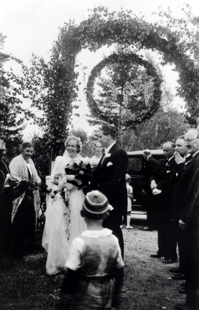 Magda Brunnberg och Carl Otto "Cotto" Glassel vid bröllopsfesten i Björkhaga.  Ovanför grinden har familjen satt upp en äreport och en krans med  brudparets initialer (någon illvillig person ansåg att bokstäverna MC var reklam för Mjölkcentralen). Runt brudparet syns på vänster sida okänd och Josefina Lundqvist.  Pojken i mössa är brudens systerson Björn Pettersson.  Mellan brudparet står Astrid Rosenius.  Vid bilen står okänd pojke och brudens svåger Georg Pettersson.  T h om brudparet okänd, med cigarrett Anders Rosenius och Georg Petterssons far som var droskägare samt okänd