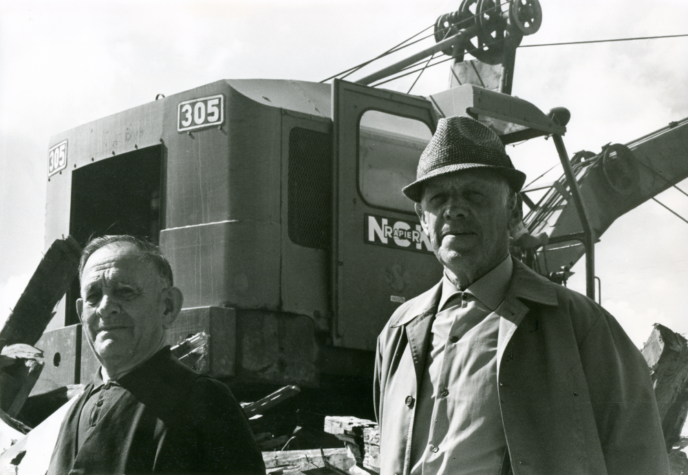 Rivning av Vallentuna stationshus pågår. Före detta lokförare E Österberg och Helge Almqvist är på plats vid stationen. I bakgrunden syns grävskopan.
•Fotografiet ingår i bildserien ”Vardagsliv i Vallentuna kommun” av Rachael Gough-Azmier & Gunilla Blomé, Haga Studios HB.