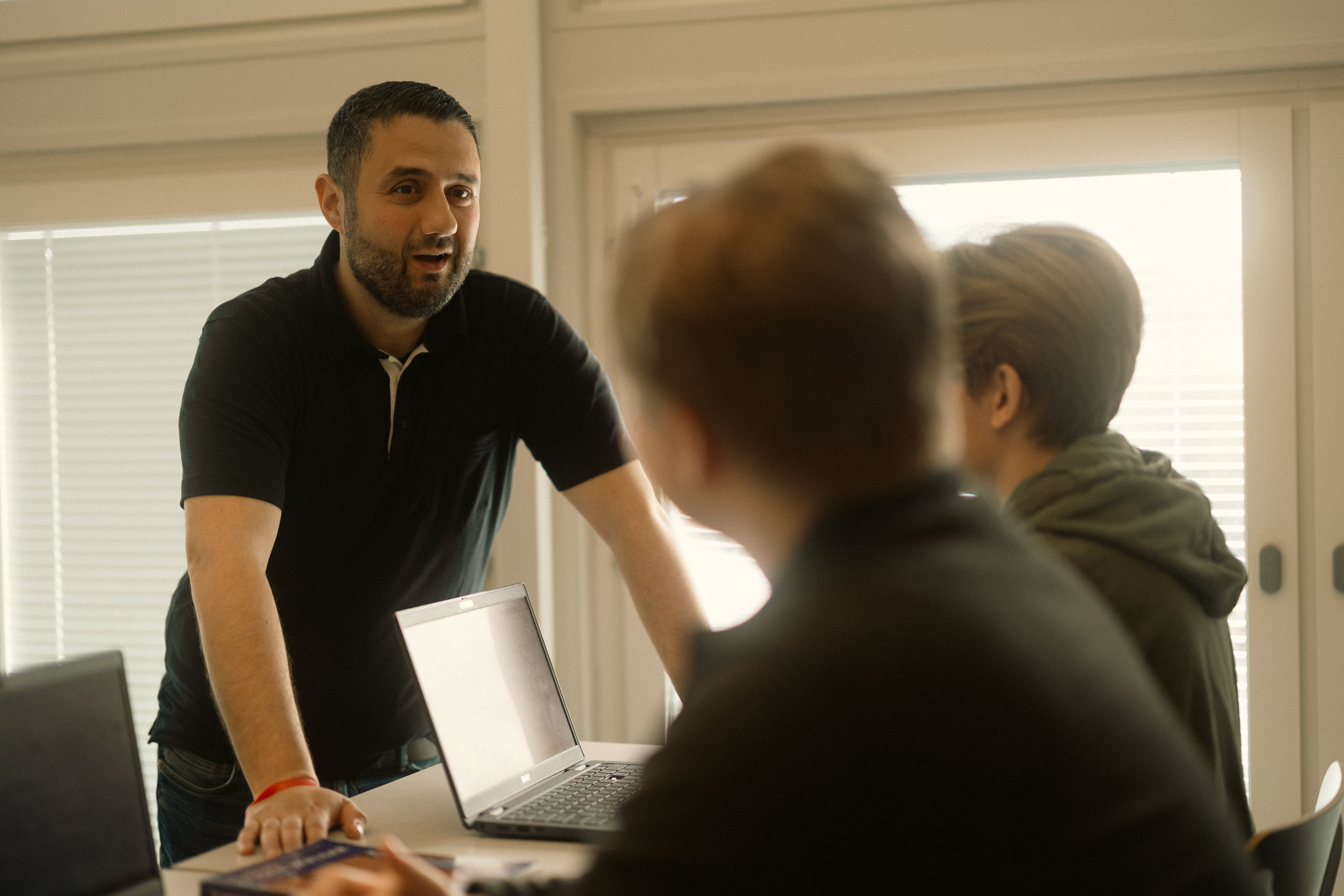 Lärare pratar med två elever som sitter intill ett bord på sina skoldatorer.