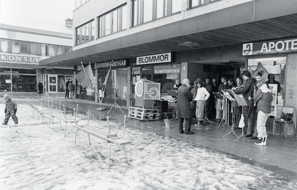 Sveriges arbetareparti m fl socialistiska partier i Vallentuna ordnar första maj-demonstration med samling bakom Centrumbio för att tåga till Tuna torg. Här har man kommit fram till Tuna torg där en musikorkester samlats. En talarstol är ännu tom. Det är snöglop och mycket folktomt på Tuna torg