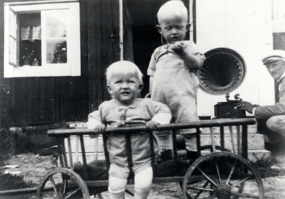 Tvillingarna Einar och Erik Eliasson, födda 1922, leker i en skrinda utanför Hjälmsättratorp i Österåker.  Fadern Einar Eliasson sitter på huk vid trappan i bakgrunden.  På trappan står en vevgrammofon med stor tratt