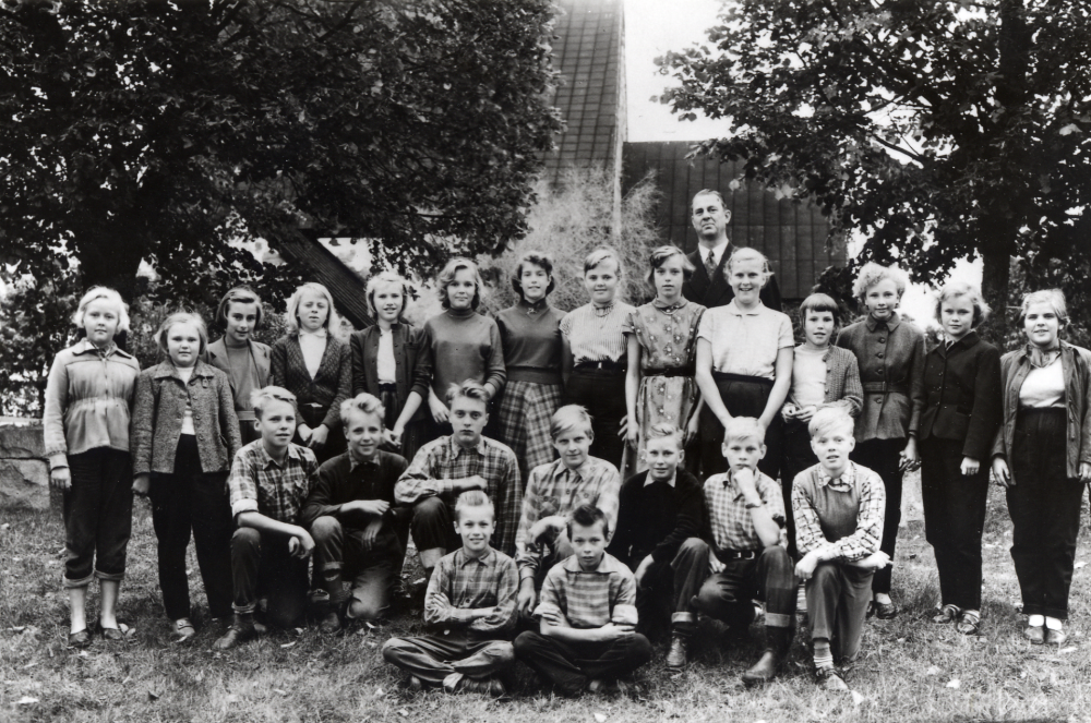 Läraren Gösta Sahlin tillsammans med skolbarnen i Össeby-Garns kyrkskola framför Össeby-Garns kyrka läsåret 1955-56.  I bakre raden står fr v Lisbet Johansson, Marianne Gustavsson, Sonja Fredriksson, Solveig Karlsson, Kristina Stenberg, Eva Nilsson, Barbro Öhman, okänd, Barbro Tillman, Monika Johansson, Gunnel Eriksson, Ann-Mari Karlsson, Ulla Andersson och Lille-Mor Öberg.  På knä står fr v Lars-Åke Nilsson, Bengt Nilsson, Sune Gustavsson, Arne Gustavsson, Sune Karlsson, Owe Jansson och Ove Söderblom.  Sittande längst fram K-G Wettervik och Lasse Jansson
