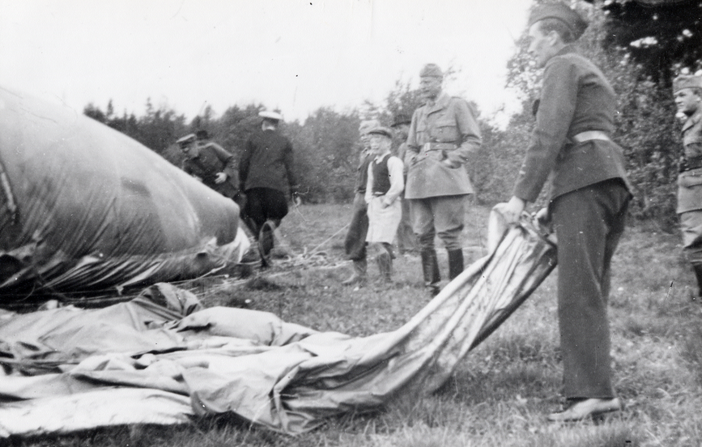 En spärrballong från England har landat vid Hall i Kårsta. En mängd män har samlats kring ballongen. Polis och militär personal finns på plats liksom åskådare, enbart män och småpojkar