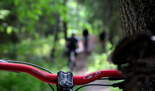 Ett MTB-styre i skogen. Andra cyklister syns suddiga i bakgrunden.