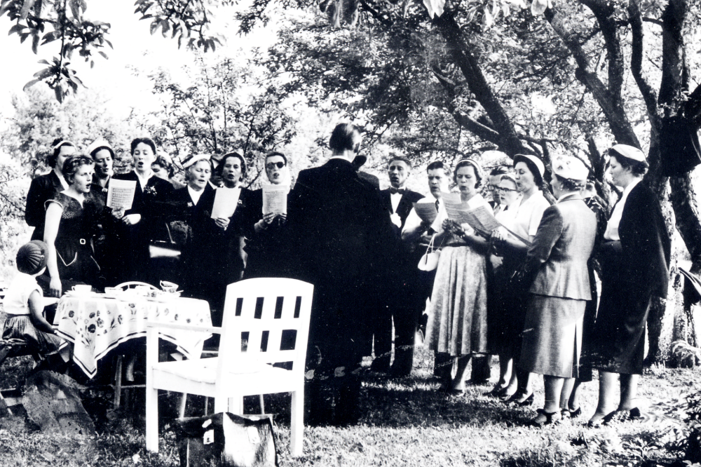 Vallentuna kyrkokör sjunger under ledning av kantor Holger Hovmark i Skärfelten vid Uppsala-Näs kyrka. Fr v nr 1 Eva Gustavsson (Anna-Lisa Gustavssons dotter), nr 2 ?, nr 3 ?, nr 4 Majken Jansson, nr 5 Evy Lööv, nr 6 Olga Jansson, nr 7 Aina Bornholm, nr 8 Bengta Jonsson, nr 9 Holger Hovmark, nr 10 Martin Nilsson, nr 11 Gösta Bornholm, nr 12 Elsie Bohman, nr 13 Erik Svenander, nr 14 ?, nr 15 Dagny Westling, nr 16 Maja Sundborg, nr 17 Olga Jonsson och nr 17 Anna Fougman??. Körmedlem Aina Bornholms mamma boddde i Skärfelten