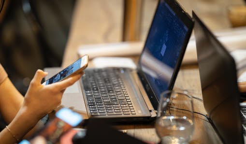 En person som sitter vid en laptop och håller i en mobiltelefon