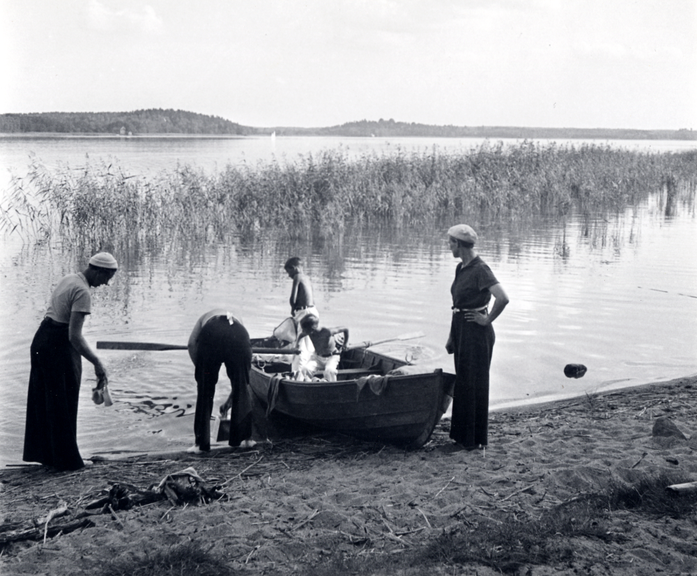 Strandhugg vid Vallentunasjön. Familjen Andersson har lagt till med roddbåt vid Vallentunasjöns norra strand t v om badbryggan. Längst t v Margareta Andersson Josef Andersson med ryggen mot kameran, i båten sitter sönerna Svante Andersson och Jerker Andersson och vid fören står Margaretas syster Elsa Olsson Fotograferat med självutlösare