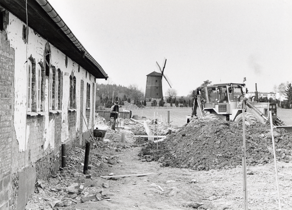 Vallentuna kyrkogård utökas mot väster.  Här arbetar en schaktmaskin vid prästgårdens ladugård.  I bildens mitt ligger Väsby kvarn