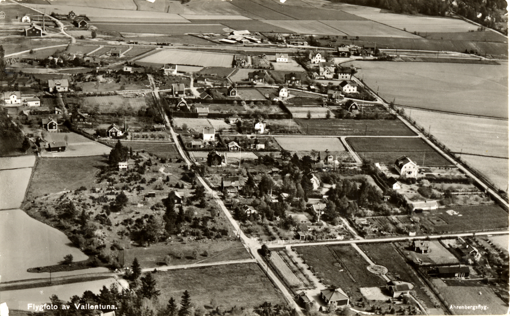 Flygfoto över Vallentunas centrala delar. Längst upp i vänstra hörnet ligger Vallentuna tegelbruk. Allévägen längst t h och parallell väg t v är Källvägen. Längst ner vid Allévägens början ligger Vallentuna järnvägsstation och järnvägshotellet Flugan.  Järnvägen är ännu inte elektrifierad och motorvagnsstallet vid Åby gata  byggs först 1938-39