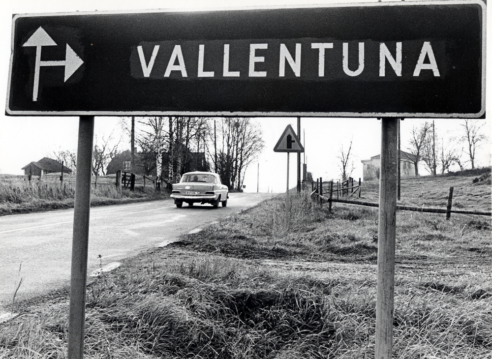 Gamla Norrtäljevägen i Angarn. Fotograferat genom avfartsskylten mot Vallentuna. På vänster sida om vägen ligger Angarns skola och på vägen kör en Mercedes