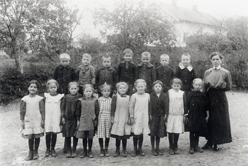 Lärarinnan Edit Lundgren, dotter till skolläraren Lundgren, tillsammans med skolbarnen utanför Vallentuna småskola, i skolhuset fanns senare bl a pastorsexpedition.  I bakgrunden Klockargården. I främre raden nr 3 fr v står Edit Carlsson Stora Ekeby gård. I småskolan gick 2 klasser enligt påskrift på kortets baksida skrivet av Edits bror Ejnar Carlsson