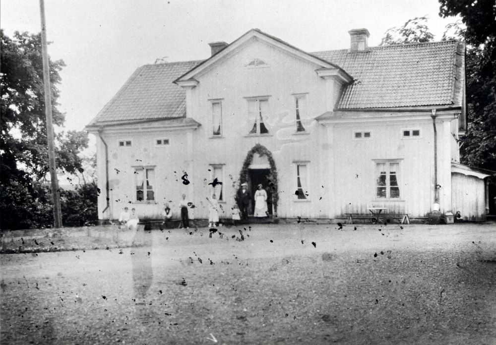 Kusta gård. Dörren är lövad med en girlang. Åtta personer står framför huvudbyggnaden.
Fotografiet fläckat av bläckskrift