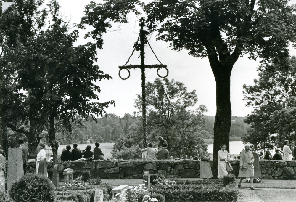 Traditionsenligt midsommarfirande på Kyrkängen i Vallentuna. Midsommarstången är rest, fotograferad från kyrkogården.
•Fotografiet ingår i bildserien ”Vardagsliv i Vallentuna kommun” av Rachael Gough-Azmier & Gunilla Blomé, Haga Studios HB.