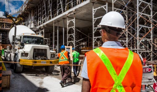 En byggarbetare står med ryggen mot kameran, tittandes på en byggarbetsplats. Byggarbetaren har en vit bygghjälm på sig.
