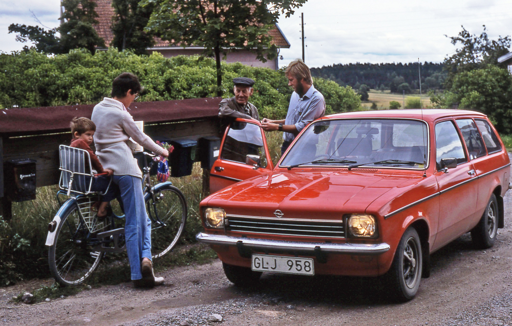 En arbetsdag med lantbrevbärare Mikael Mattson. Lantbrevbäring i Rävsta;  Ulla Johansson med sin son bak på cykeln, Gustav Andersson (bor i Nygård som ligger i skogen som skymtar bakom bilen) och lantbrevbärare Mikael Mattsson. I huset, i bakgrunden, bor Karin Jansson. Gustav Andersson var en duktig skytt och brukade hjälpa till med grävarbeten av diken.
