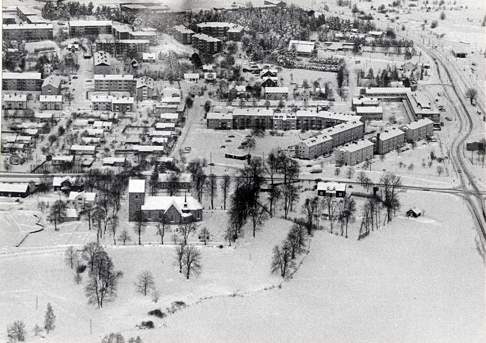 Flygfoto över Vallentuna centrala delar med Vallentuna kyrka i förgrunden. Bakom kyrkan går Väsbyvägen. Längst t h Banvägen och Roslagsbanan med Vallentuna centrum t v. Längre västerut Prästgårdsgärdet med villabebyggelse. I bakgrunden flerfamiljshus på Mörbyvägen och Fornminnesvägen, allra längst bort skymtar industriområdet vid Teknikvägen