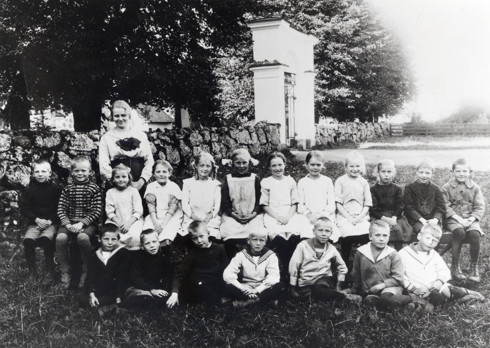 Lärarinnan Margit Oppermann tillsammans med skolbarnen utanför Vallentuna kyrka.  Bakre raden fr v Oskar Sjöstrand, Börje Johansson (Blidstam), Aina Hofström, Elsa Karlsson, Ingrid Johansson, okänd, okänd, okänd, Edit Olsson, Karin Nordström, okänd och Holger Eriksson.  Nedre raden fr v Sven Håkansson, Karl Sjöstrand, Ragnar Pettersson, Arne Lindström, Sixten Mörk, Gösta Andersson och Stig Henningsson.  Förmodligen hade barnen sitt klassrum i Klockargården och detta var Margit Oppermanns första klass