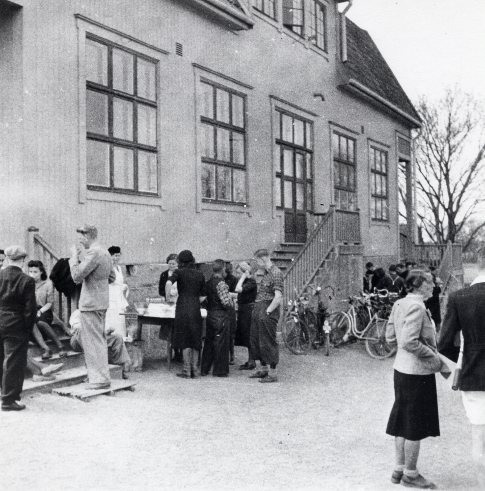 Vita eller Gula skolan, Hjälmsta.  På skolgården pågår servering, försäljning eller basar, kanske för skolresa, frivilliga brandkåren eller Röda korset.  En kvinna i vitt förkläde står vid ett bord mellan trapporna.  Kvinnan i ljus kavaj och mörk kjol t h är lärarinnan Edit Lindström.  Hon talar med en man i ljusa kortbyxor och med en tennisracket under armen.  Pågår möjigen tennisturnering vid Hjälmsta skola?