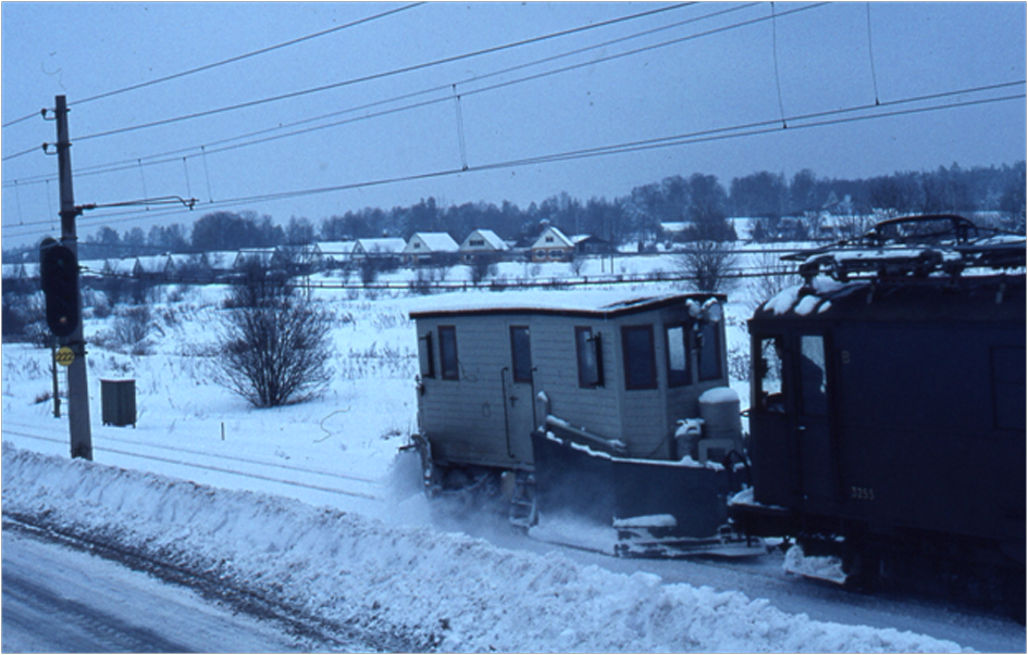 Snöplogen, påskjuten av lok 3255, på väg norrut från Vallentuna.  Tåget har just passerat Åby gata.  I bakgrunden åt vänster syns raden av kedjehus utmed Björkhagsvägen