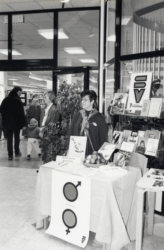 Bokfest 1996 i Vallentuna bibliotek med tema barnkultur.  I foajén  presenterar Studieförbundet Vuxenskolan sin verksamhet, t v i dörren står Inger Kjellerby-Åberg och vid bordet Anne Spetz