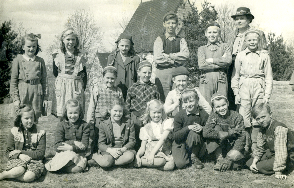 Lärarinnan Ingrid Lindell tillsammans med skolbarnen i klasserna 3-4 utanför Össeby-Garns kyrkskola.  I bakgrunden skymtar Össeby-Garns kyrka.  Stående fr v Gunilla Wettervik (Holm) Brottby, Ragna Alstermark Ösby, Berit Hammarström Svista, Harry Bergqvist, Lasse Rödin, lärarinnan och Lasse Gustavsson Sjötorp.  Knästående i mitten fr v Björn Larsson, Hasse Stjernholm och Gösta Johansson Hakunge.  Sittande fr v Eva Britt Persson (Segerman), Astrid Gustavsson (Johansson) Skeppartorp, Britt Nordström (Birgersson) Lövhamra, Ulla Johansson (Lundqvist) Ösby, Gösta Andersson Pälsen, Stig Jansson Ösby och Rune Johansson Gillberga