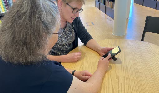 Två kvinnor tittar på en telefonskärm, den ena instruerar den andra genom att peka på skärmen.