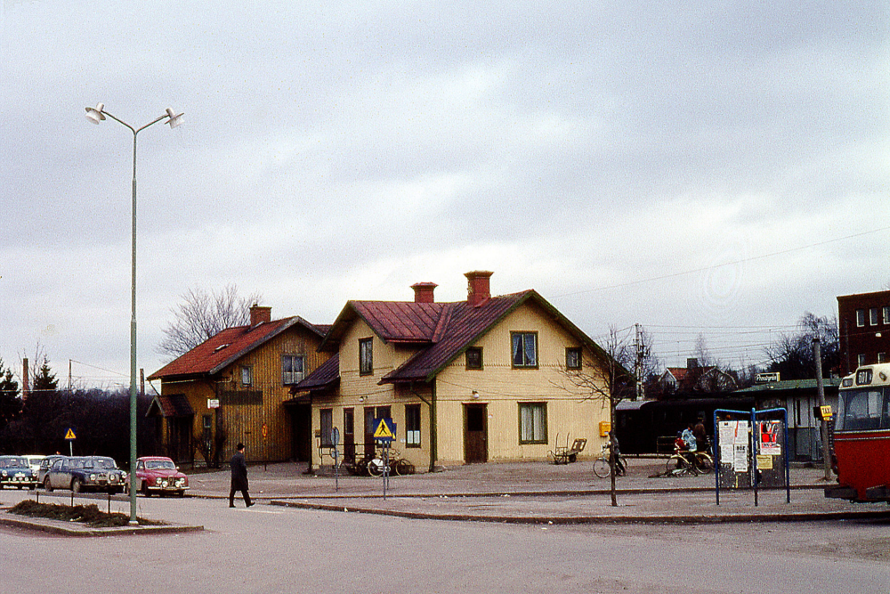 Det orangefärgade huset bakom den gula stationsbyggnaden är det före detta järnvägshotellet med restaurangen som kallades för Flugan. Längst till vänster i bilden syns skorstenarna till de båda tegelbruken. Till höger om stationshuset syns Pressbyråns kiosk. På andra sidan järnvägen syns hörnet på Tellus maskins huvudbyggnad. Färgbild