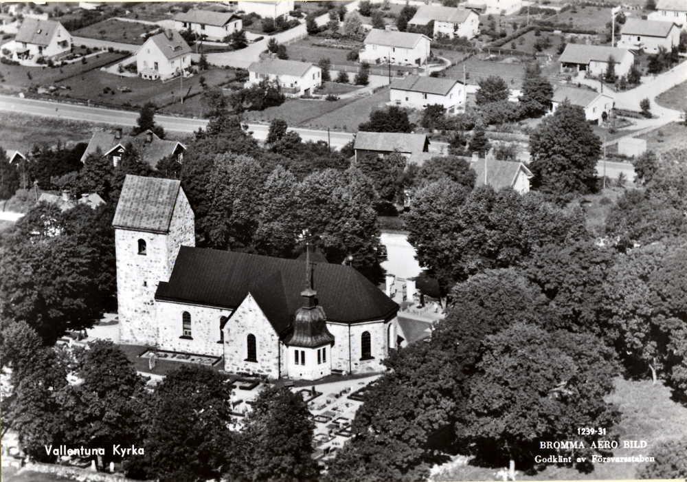 Flygfoto över Vallentuna kyrka med enfamiljshus på Prästgårdsgärdet. Mellan kyrkan och bostadsområdet går Väsbyvägen