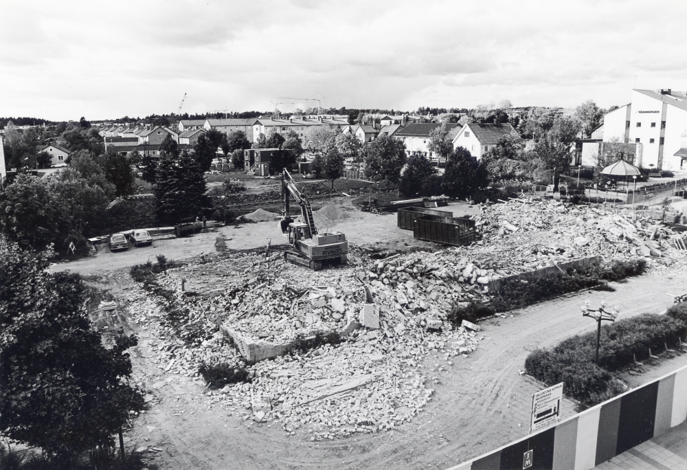 Vallentuna kommunalhus rivs i september 1985. På dess plats skall ett nytt och mer ändamålsenligt kommunalhus byggas.  T h syns Tärningen med Paviljong och byggkrananr i bakgrunden visar på bygget av flerfamiljshus med bl a servicelägenheter i kvarteret Sommarvägen, Källvägen, Tvärvägen och Allevägen