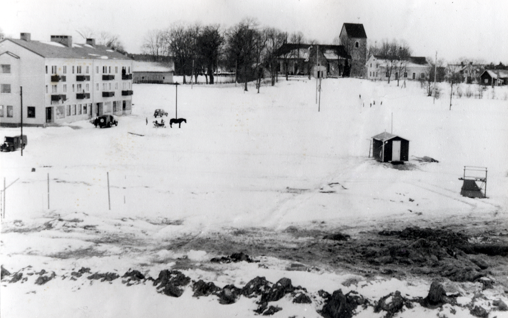 Prästgårdgärdet  där husen vid  Skördevägen  (i förgrunden) ännu ej byggts. Till vänster flerfamiljshusen längs  Centralvägen, här bebyggt endast på östra sidan, med affärslokaler i bottenplan. Några bilar står parkerade och en häst drar en släde. Enligt den ursprungliga planen skulle gärdet bebyggas endast med enfamiljshus eftersom markförhållandena var mindre lämpade för stora byggnader. I bakgrunden  Vallentuna kyrka.