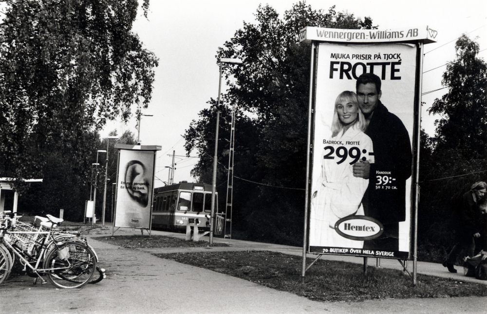 Bällsta järnvägsstation med reklampelare för Hemtex frotté.  Ett blått tåg är på väg in på plattformen