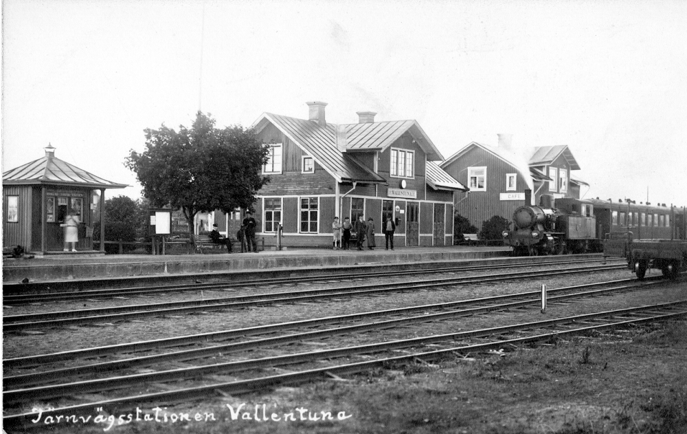 Vallentuna järnvägsstation och Järnvägsrestaurangen Flugan,  Flugan har en skylt med Café på gaveln. Söder om stationshuset syns Ester Gustavsson kiosk med omgjort tak i pagodliknande stil som kan jämföras med kiosktak på bl. a. Tegelbacken i Stockholm. På plattformen finns både järnvägspersonal och människor.  På spår 1 står ett ånglok med en passargerarvagn.  Semaforen saknas på bilden