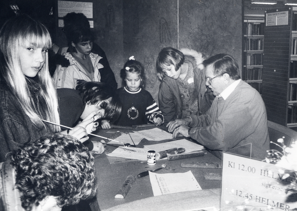 Bokfest 1991 i Vallentuna bibliotek. Boken Runstenar i Vallentuna av Helmer Gustavson förstadagssäljs.  Vid samma bord hjälper Erold Sandqvist, Vallentuna biblioteks vänner, barn att göra en runstav