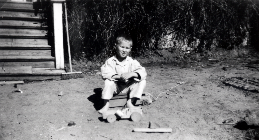 Timo Suomalainen leker vid kökstrappan på Ekskogens gård med bilar av trä.  T h syns delar av en ladugård byggd av stenar och grankottar  Vid kortet i albumet står antecknat "God morgon"

Timo och hans lillebror Sami kom till Sverige som finska krigsbarn 1944. Timo adopterades senare av ägarparet på Ekskogens gård i Kårsta, Inez och Manfred Ekholm. Läs mer i Timo Ekholms bok Krigsbarn 40.